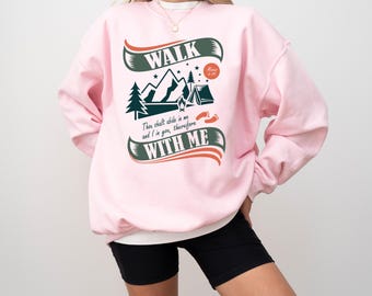 Könnte beinhalten: Hellrosa Sweatshirt mit Camping-Motiv. Das Design zeigt die Worte "WALK" und "WITH ME", sowie eine Berglandschaft, Bäume und ein Zelt. Das Sweatshirt hat lange Ärmel.