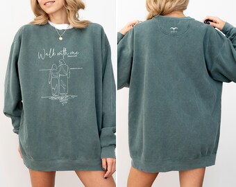 Könnte beinhalten: Ein teal-farbenes Sweatshirt mit dem Schriftzug "Walk with me" und einer Strichzeichnung von zwei Figuren. Auf der Rückseite des Sweatshirts befindet sich ein kleines weißes Logo. Das Sweatshirt hat eine lockere Passform.