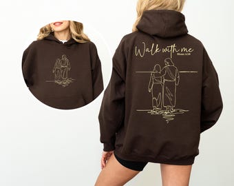 Könnte beinhalten: Ein dunkelbrauner Kapuzenpullover mit Kängurutasche. Auf der Rückseite befindet sich eine goldene Strichzeichnung von zwei Figuren mit dem Text "Walk with me Moses 6:34". Auf der Vorderseite befindet sich eine kleinere Version derselben Zeichnung.