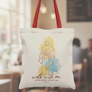 May include: A white tote bag with red handles. The bag features a line drawing of two figures with watercolor accents in yellow, blue, and peach. The words "walk with me" and a Bible verse are printed in brown.