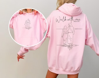 Könnte beinhalten: Hellrosa Kapuzenpullover mit einer schwarzen Strichzeichnung von zwei zusammengehenden Figuren. Auf der Rückseite des Hoodies steht der Text "Walk with me" und "Moses 6:34". Eine kleinere Version der Zeichnung befindet sich auf der Vorderseite.