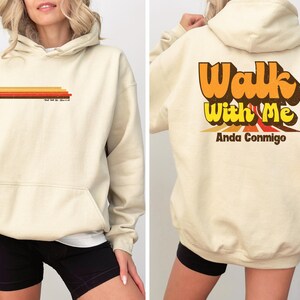 May include: Cream-colored hoodie with a front pocket and drawstring hood. The front features a horizontal design of orange, red, and brown stripes, with text below. The back displays the words "Walk With Me" in orange and yellow, with "Anda Conmigo" below.