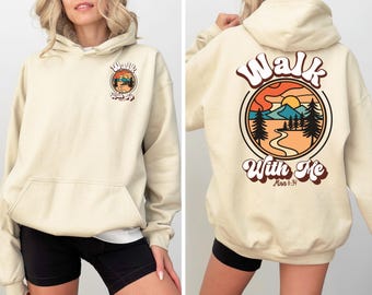 Könnte beinhalten: Cremefarbener Kapuzenpullover mit Fronttasche und Kapuze. Auf der Vorderseite befindet sich eine kreisförmige Grafik mit dem Text "Walk With Me". Auf der Rückseite des Hoodies ist eine größere Version der Grafik mit einer Berglandschaft und demselben Text abgebildet.