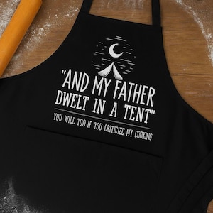 May include: Black apron with white text: "AND MY FATHER DWELT IN A TENT" and "YOU WILL TOO IF YOU CRITICIZE MY COOKING". Features a tent and moon graphic. Includes a pocket. Rolling pin and heart-shaped cookie cutters are in the background.