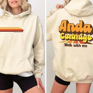 Może przedstawiać: Kremowa bluza z kapturem z przednią kieszenią i kapturem. Przód posiada poziome paski w odcieniach pomarańczu i brązu, z tekstem poniżej. Tył wyświetla słowa "Anda Conmigo" i "Walk with me" w stylu retro.