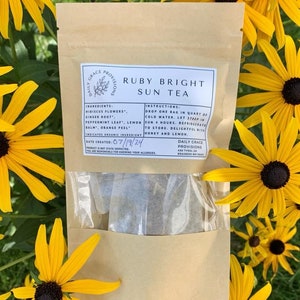 May include: A brown paper bag with a clear window showing a tea bag inside. The bag has the text "Daily Grace Provisions" and "Ruby Bright Sun Tea" printed on it. The bag is surrounded by yellow flowers with dark centers.