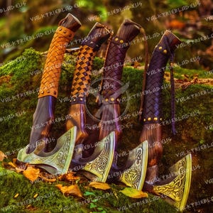 May include: Four hand-crafted axes with leather wrapped handles. The axes have intricate metal designs on the blades and are displayed on a mossy rock.