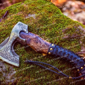 Könnte beinhalten: Eine silberne Axt mit einem Holzstiel, der in schwarzes Leder mit Messingnieten gewickelt ist. Der Axtkopf hat komplizierte Knotenmuster.