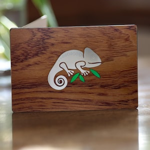 May include: A rectangular wooden box with a detailed wood grain pattern. A silver chameleon design with green leaves is affixed to the front. The chameleon is perched on a branch, with its tail curled. The box is likely used for storage or display.