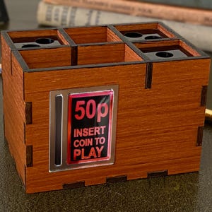 May include: A wooden desk tidy with a retro arcade game theme. The organiser has multiple compartments and a slot that reads "50p INSERT COIN TO PLAY" in red and white. The wood has a warm, reddish-brown tone.