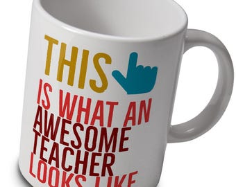 May include: White ceramic mug with the text "THIS IS WHAT AN AWESOME TEACHER LOOKS LIKE" in yellow, red, and blue. The mug has a curved handle and a blue hand pointing upwards. A gift for teachers.