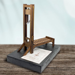 May include: A wooden model of a guillotine on a black slate base. The base has a printed card with the text "Madame Guillotine".