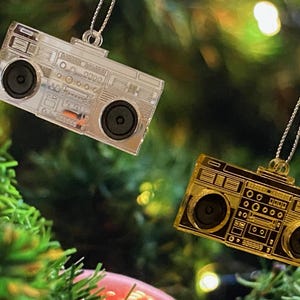 May include: Two silver and gold boombox baubles hanging from a Christmas tree. The baubles are shaped like vintage cassette players with buttons and dials.