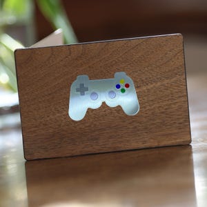 May include: A rectangular wooden plaque featuring a silver-coloured video game controller design. The controller has a plus sign, two circles, and four coloured buttons. The wood has a natural grain.