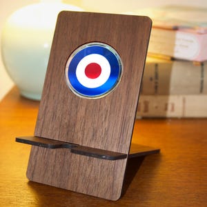 May include: A wooden phone stand with a black phone holder. The stand has a round, blue, white, and red target design on the front.