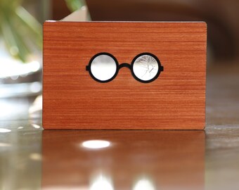 May include: A wooden card with a pair of black-rimmed glasses with cracked lenses. The card has a wood grain pattern.