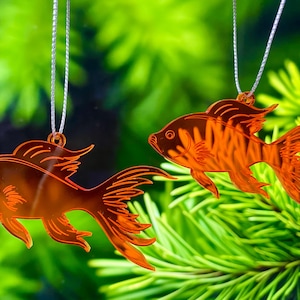 Peut inclure: Deux ornements en forme de poisson orange suspendus par des fils argentés. Les ornements présentent des motifs détaillés d'ailerons et de queue. Le fond est vert flou, suggérant un thème naturel ou aquatique. Ce sont probablement des objets décoratifs.