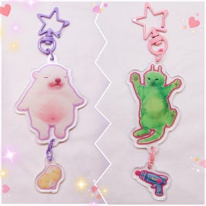 May include: Two acrylic keychains. One features a white bear with pink accents, a purple star-shaped clasp, and a small potato charm. The other shows a green alien with a pink star clasp and a ray gun charm.