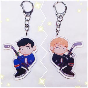 May include: Two acrylic hockey player keychains. One keychain features a player in a blue uniform, the other in a black uniform. Both keychains have a silver clasp and a hockey stick.