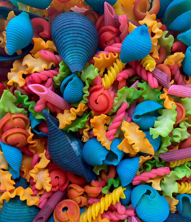 Puede incluir: Una vibrante variedad de formas de pasta coloridas, incluyendo conchas, espirales y penne. La pasta est&aacute; te&ntilde;ida en una variedad de colores brillantes como azul, rojo, amarillo, naranja y verde, creando una exhibici&oacute;n visualmente atractiva. Primer plano de la pasta.