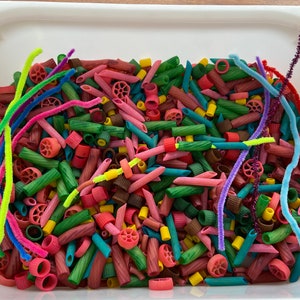 Puede incluir: Una bandeja blanca llena de formas de pasta coloridas y limpiapipas. La pasta está teñida en tonos de rosa, verde, marrón y amarillo. Limpiapipas de colores brillantes en varios colores están esparcidos por la pasta.
