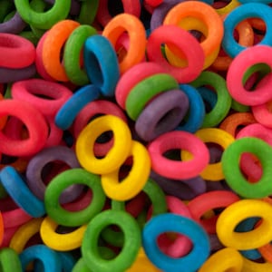 Puede incluir: Primer plano de cereales de colores en forma de anillos. La imagen está llena de una variedad de colores brillantes, incluyendo rojo, naranja, amarillo, verde, azul y morado. Los cereales están dispersos y superpuestos, creando una textura vibrante y juguetona.