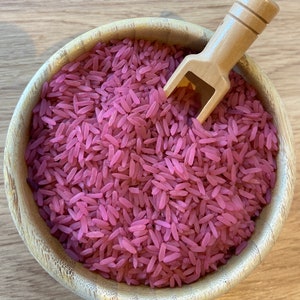 Puede incluir: Un cuenco de madera lleno de arroz rosa y una pequeña cuchara de madera. El arroz es de un color rosa vibrante, y el cuenco tiene una veta de madera natural. La cuchara está en el arroz, lo que sugiere que está listo para servir.