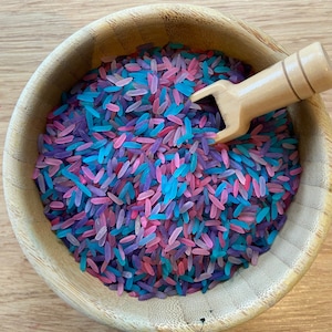 Puede incluir: Un cuenco de madera lleno de arroz teñido de colores en tonos rosa, morado y azul. Una pequeña cuchara de madera descansa en el arroz. El cuenco está sobre una superficie de madera clara. Este artículo de juego sensorial es ideal para niños.