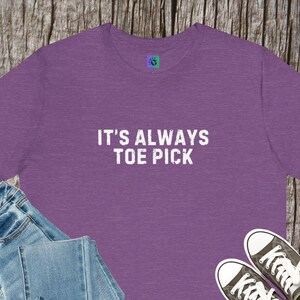 May include: A purple t-shirt with the text "IT'S ALWAYS TOE PICK" in white. The shirt is paired with blue jeans and black and white sneakers. The shirt has a small logo on the upper left.