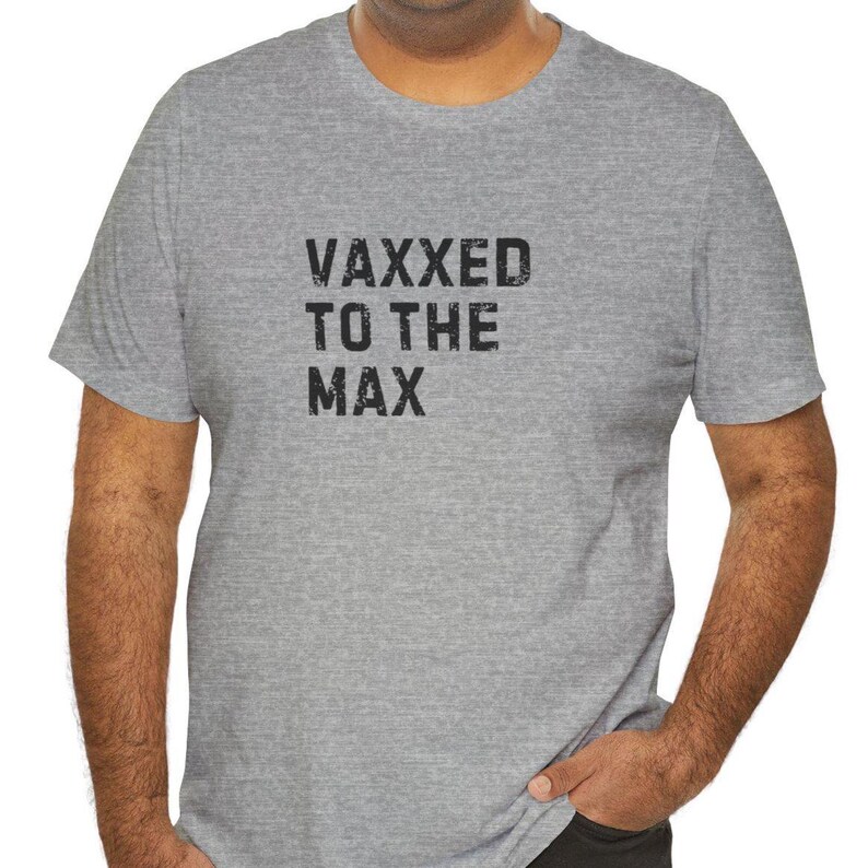 May include: A gray t-shirt with the text "VAXXED TO THE MAX" printed in black. The shirt is short-sleeved and has a crew neck. The text is in a bold, slightly distressed font.
