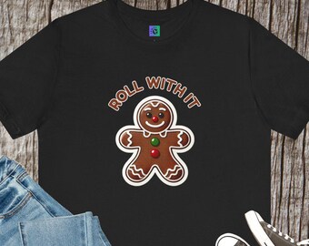 May include: Black t-shirt with a gingerbread man graphic and the text "ROLL WITH IT". The gingerbread man has a smiling face, red and green buttons, and a white outline. The shirt is paired with blue jeans and black trainers.
