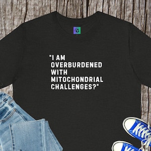 May include: Black t-shirt with the text "I AM OVERBURDENED WITH MITOCHONDRIAL CHALLENGES?" in white. A small logo is on the upper right. Blue jeans and blue trainers are partially visible in the image.