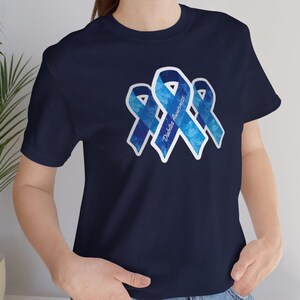 May include: Navy blue t-shirt featuring three blue awareness ribbons with the text "Diabetes Awareness". The ribbons have a textured, watercolor-like appearance and are outlined in white. The shirt is a solid color.