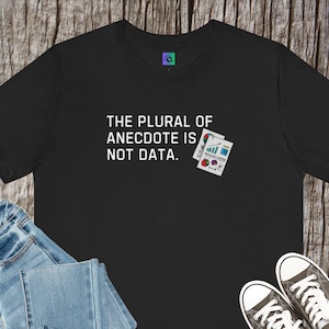 Könnte beinhalten: Schwarzes T-Shirt mit dem Text "THE PLURAL OF ANECDOTE IS NOT DATA." Eine kleine Grafik von zwei Dokumenten mit Diagrammen ist neben dem Text aufgedruckt. Das Shirt wird mit blauen Jeans und Turnschuhen kombiniert.