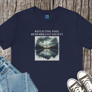 May include: Navy blue t-shirt featuring a graphic of the Washington Monument reflected in a pool under a cloudy sky. The text "REFLECTING POOL REMEMBRANCE SOCIETY" is printed above the graphic. The shirt is paired with blue jeans and black sneakers.
