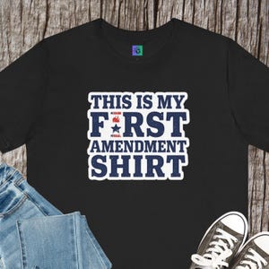 Può includere: T-shirt nera con la scritta "THIS IS MY FIRST AMENDMENT SHIRT" in bianco e blu. L'immagine mostra la maglietta con jeans blu e scarpe da ginnastica bianche e nere, su uno sfondo di legno.