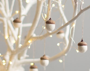 May include: A white branch tree with white lights and brown felt acorn ornaments hanging from the branches. A white ceramic tree is in the foreground.