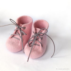 May include: Two pairs of pink felt baby booties with gray laces. The booties are tied with a bow and are on a white background.