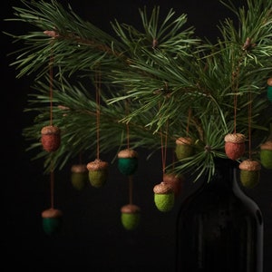 Peut inclure: Une branche de pin avec des glands en feutre vert suspendus dans un vase noir. Les glands sont de différentes nuances de vert, de brun et de rouge.