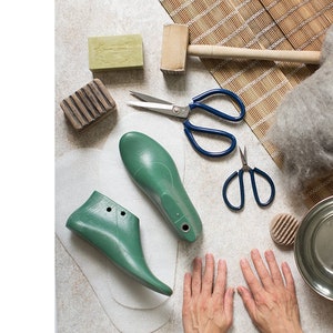 May include: A flat lay of supplies for making felt slippers, including two green shoe forms, a ball of gray wool, two pairs of scissors, a wooden mallet, a bar of soap, a wooden scrubber, and a metal bowl.