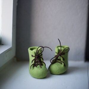 May include: A pair of green felt booties with brown laces. The booties are tied and sitting on a white surface.