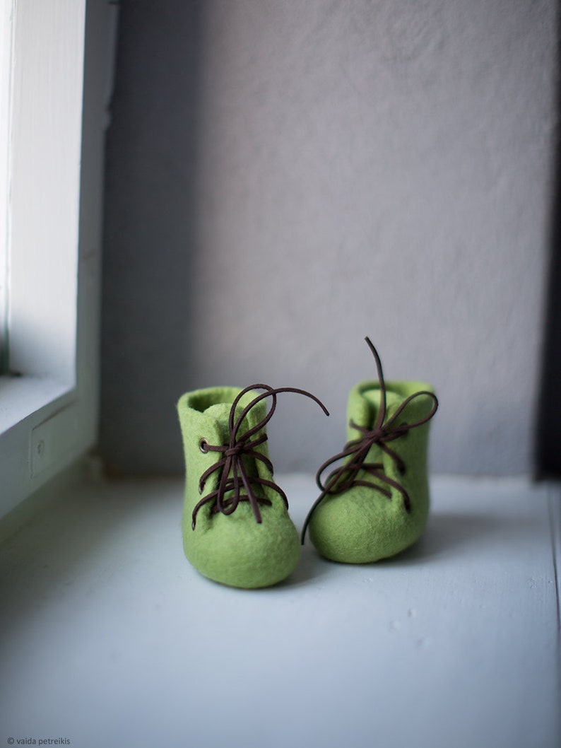 May include: A pair of green felt booties with brown laces. The booties are tied and sitting on a white surface.