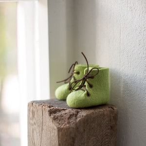 May include: A pair of green felt baby booties with brown laces. The booties are sitting on a wooden block.