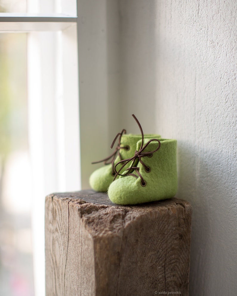 May include: A pair of green felt baby booties with brown laces. The booties are sitting on a wooden block.