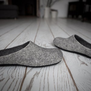 May include: A pair of gray felt slippers on a white wooden floor. The slippers are simple and comfortable, with a rounded toe and a flat sole.