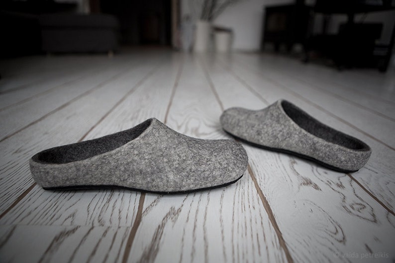 May include: A pair of gray felt slippers on a white wooden floor. The slippers are simple and comfortable, with a rounded toe and a flat sole.