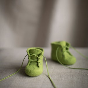 May include: Two pairs of green felt baby booties with laces. The booties are on a light brown linen surface.