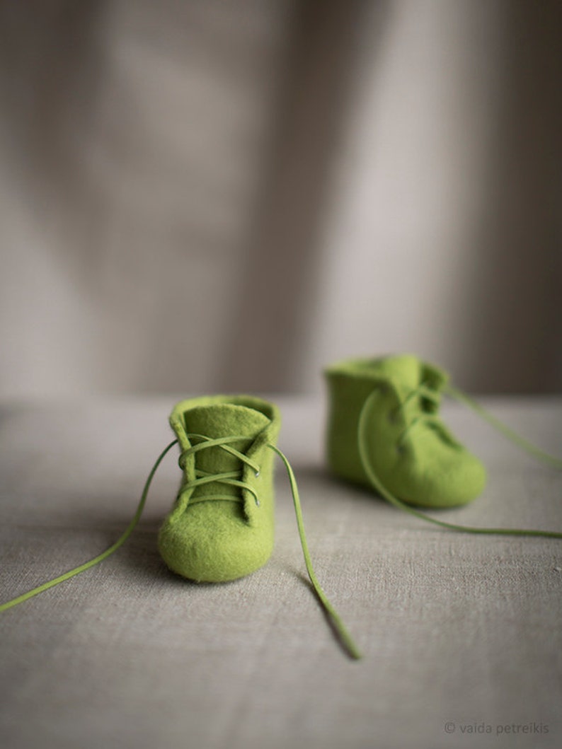 May include: Two pairs of green felt baby booties with laces. The booties are on a light brown linen surface.