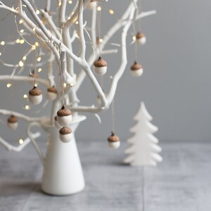 Pu&ograve; includere: Un albero di rami bianchi dipinto con un vaso a forma di brocca bianco e ornamenti di ghianda marroni appesi ai rami. L'albero &egrave; decorato con luci fatate bianche. Un albero di legno bianco ritagliato &egrave; in primo piano.