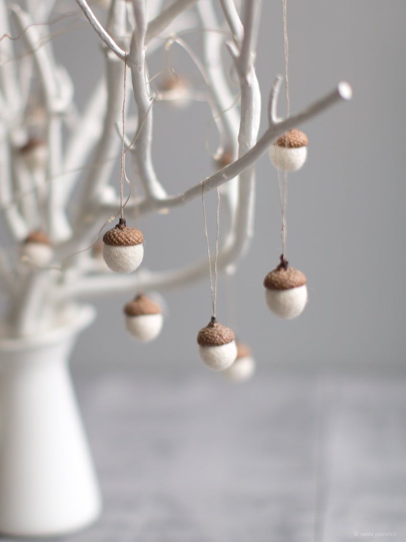 Pu&ograve; includere: Un ramo bianco con ornamenti a forma di ghianda in feltro marrone appesi ad esso. Il ramo &egrave; in un vaso bianco su una superficie grigia.
